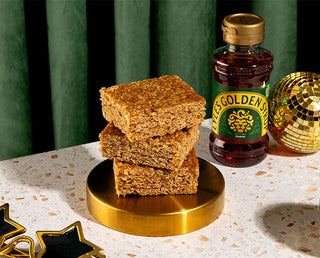 Three squares of flapjacks stacked on a gold plate, next to a Lyle's Golden Syrup Pouring Bottle and gold disco balls nearby.