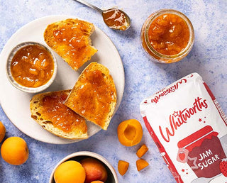 Toast slices topped with homemade apricot jam, next to a freshly made jar of jam, fresh apricots, and a Whitworths Jam Sugar bag.