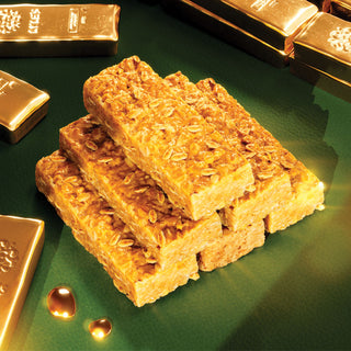 A stack of Lyle's Golden Syrup Flapjacks on a green surface with shiny gold bars in the background.