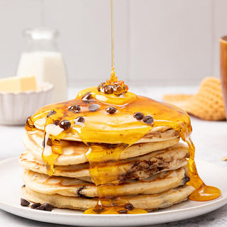 Stack of pancakes drizzled with Lyle's Golden Syrup and topped with chocolate chips, next to a Lyle's Golden Syrup 700g Pouring Bottle with milk and butter in the background.