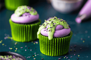 Two Halloween cupcakes with green and purple frosting on a dark surface.