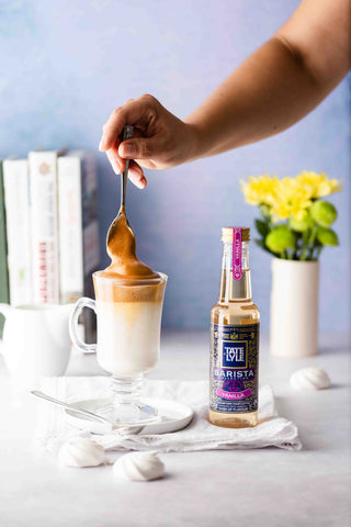 A person stirring whipped coffee in a glass cup next to a Tate & Lyle vanilla syrup bottle.