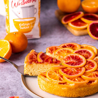A vibrant orange upside-down cake with caramelized blood orange slices, with a slice being served. Fresh oranges sit nearby with a bag of Whitworths Caster Sugar.