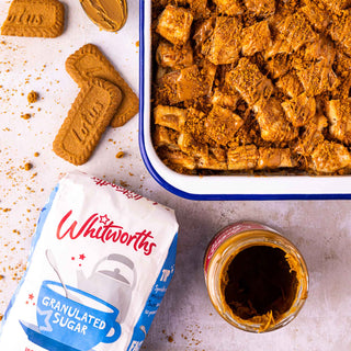 Biscoff bread and butter pudding topped with crushed biscuits, served alongside a bag of Whitworths granulated sugar and a jar of Biscoff spread.