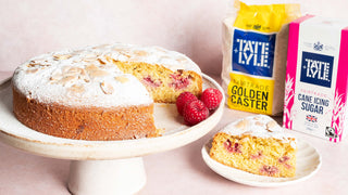 An almond and raspberry cake sits on a cake stand, with a slice placed on a nearby plate. Next to bags of Tate & Lyle Golden Caster and Icing Sugar.