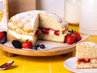 A giant scone cake filled with clotted cream and strawberry jam on a wooden cake stand, surrounded by fresh berries and baking ingredients.
