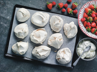 10 meringue nests on a baking tray next to a bowl of strawberries and fresh cream