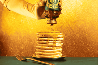 A stack of fluffy golden pancakes drizzled with Lyle’s Golden Syrup, served on a white plate for a delicious breakfast.