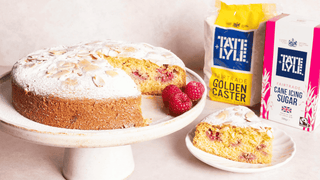 An almond and raspberry cake sits on a cake stand, with a slice placed on a nearby plate. Next to bags of Tate & Lyle Golden Caster and Icing Sugar.