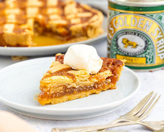 A slice of Lyle’s Easy Treacle Tart with a golden crust, topped with cream, next to a tin of Lyle's Golden Syrup 454g Tin. The full tart can be seen in the background oozing with delicious golden syrup.