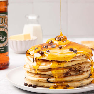 Stack of pancakes drizzled with Lyle's Golden Syrup and topped with chocolate chips, next to a Lyle's Golden Syrup 700g Pouring Bottle with milk and butter in the background. 