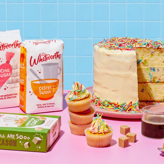 A large birthday cake sliced, next to a pile of cupcakes and a few Whitworths Sugar bags