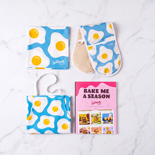 A kitchen accessory set laid out on a white marble surface, including a blue-and-white egg-pattern towel, matching oven mitt, beige foot warmer, and a pink 'BAKE ME A SEASON' recipe book. The items feature a bright blue background with white eggs and yellow yolks.