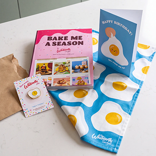 A Whitworths gift set featuring a pink 'Bake Me A Season' cookbook, a blue 'Happy Birthday' greeting card, a matching blue and white tea towel with fried egg patterns, a small egg-shaped pin on a card, and a brown paper bag, arranged on a white countertop.