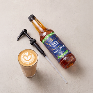 A bottle of Tate & Lyle Barista Hazelnut Syrup with a black manual pump and clear straw, displayed next to a glass of latte with leaf art on a light beige textured surface.