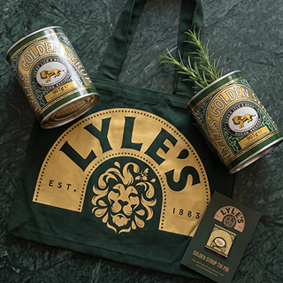 A dark green tote bag with a large gold and green Lyle's logo, two small Lyle's tins, and Lyle's Golden Syrup tin pin