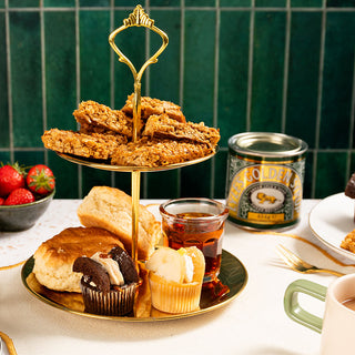 A two-tiered dessert stand with flapjacks, cupcakes, and scones. A tin of Lyle's Golden Syrup sits nearby, alongside strawberries and a cup of tea.