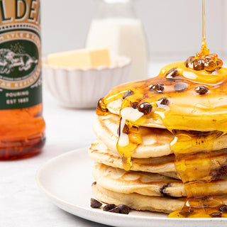 Stack of pancakes drizzled with Lyle's Golden Syrup and topped with chocolate chips, next to a Lyle's Golden Syrup 700g Pouring Bottle with milk and butter in the background.
