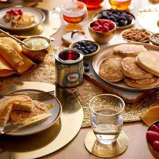 A beautifully set table featuring a stack of homemade pancakes, complemented by Lyle's Golden Syrup in both the 700g Pouring Bottle and 407g Tin, alongside fresh berries and crunchy nuts, all arranged on a golden tablecloth.