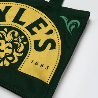 A dark green canvas tote bag featuring a large gold graphic print with the text 'STYLE'S' and '1883', along with a small green wave-like logo on the side, shown on a light gray background.
