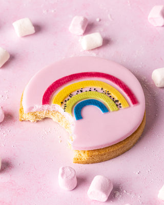 Cookie with pink icing and rainbow decoration on a pink background with marshmallows.