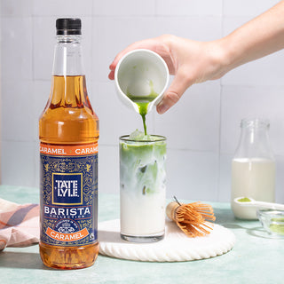 A bottle of Tate & Lyle Caramel Syrup sits beside an iced matcha latte, as matcha is gracefully poured into the glass.