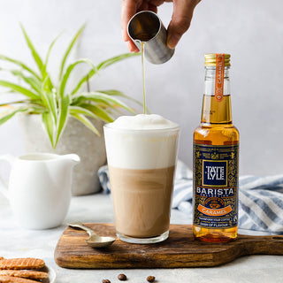 A hand pours caramel syrup from a metal jigger into a frothy latte, with a bottle of Tate Lyle caramel syrup nearby. 