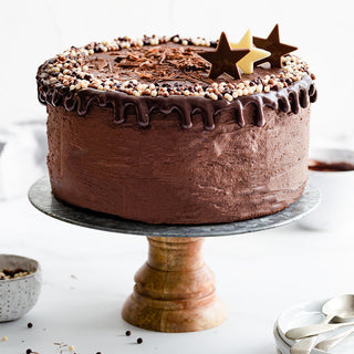 A tall iced chocolate cake on a wooden cake stand, topped with chocolate ganache, sprinkles, and star-shaped chocolates. 