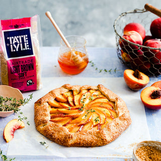 A peach galette topped with sliced peaches and thyme, with a bowl of brown sugar, honey, and peaches nearby. 