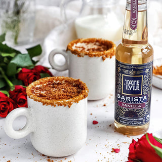 Two steaming mugs of Biscoff lattes, next to a bottle of Tate & Lyle vanilla syrup.