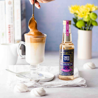 A person stirring whipped coffee in a glass cup next to a Tate & Lyle vanilla syrup bottle