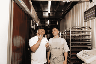Two bakers in a bakery holding a loaf of bread, with shelves and equipment in the background.