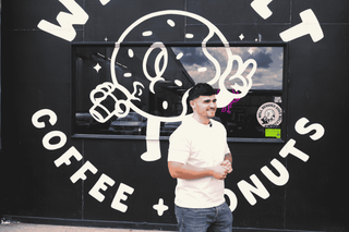 Man standing in front of a coffee and donuts shop with a large logo on the wall.