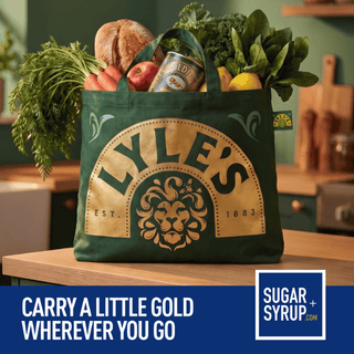 A dark green reusable tote bag with a large gold 'LYLE'S' logo and lion emblem, filled with groceries including bread, carrots, apples, kale, and lemons, set on a wooden table in a kitchen. A blue banner at the bottom reads 'CARRY A LITTLE GOLD WHEREVER YOU GO' with the Sugar + Syrup logo.