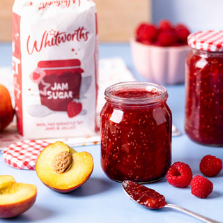 A jar of peach and raspberry jam sits alongside a spoon, surrounded by fresh raspberries and a sliced peach. In the background, a bag of Whitworths jam sugar.
